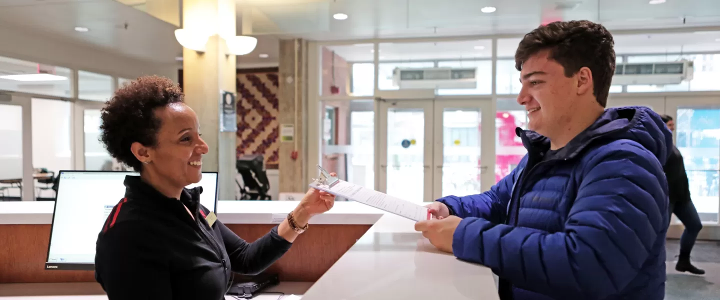 A photo of a young man handing a form to a YMCA employee at the front desk.