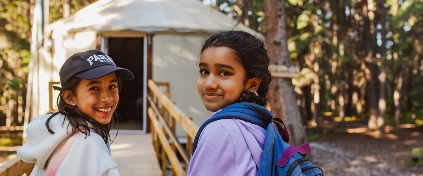 Two girls smiling in front of a yurt at camp in sunlight.