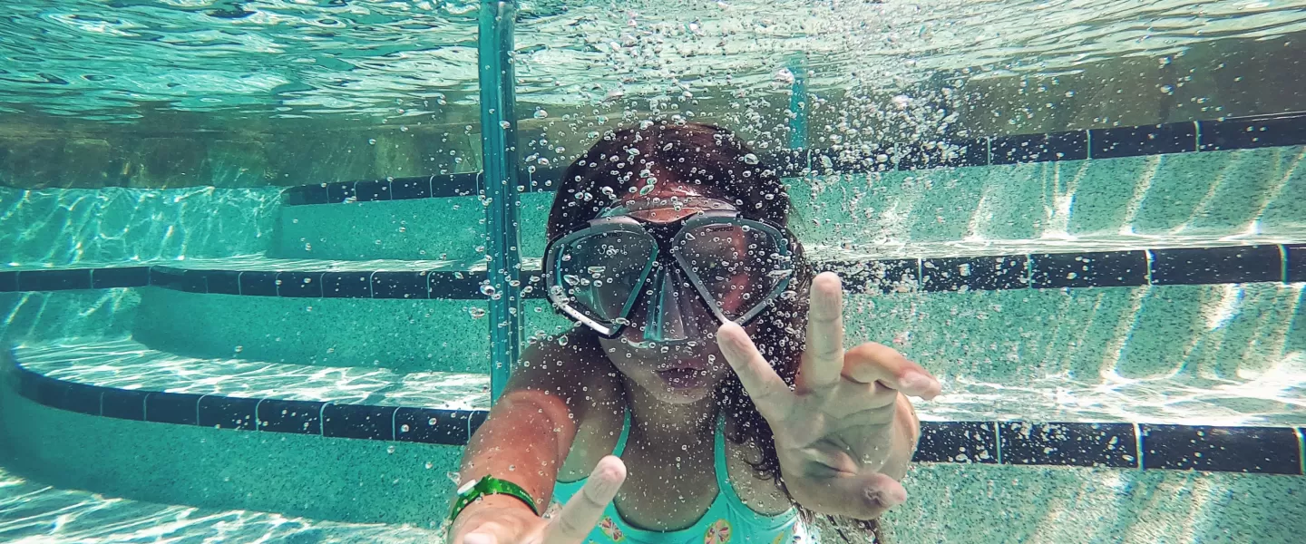 Child underwater in pool wearing snorkel mask, playfully forming peace signs with both hands.