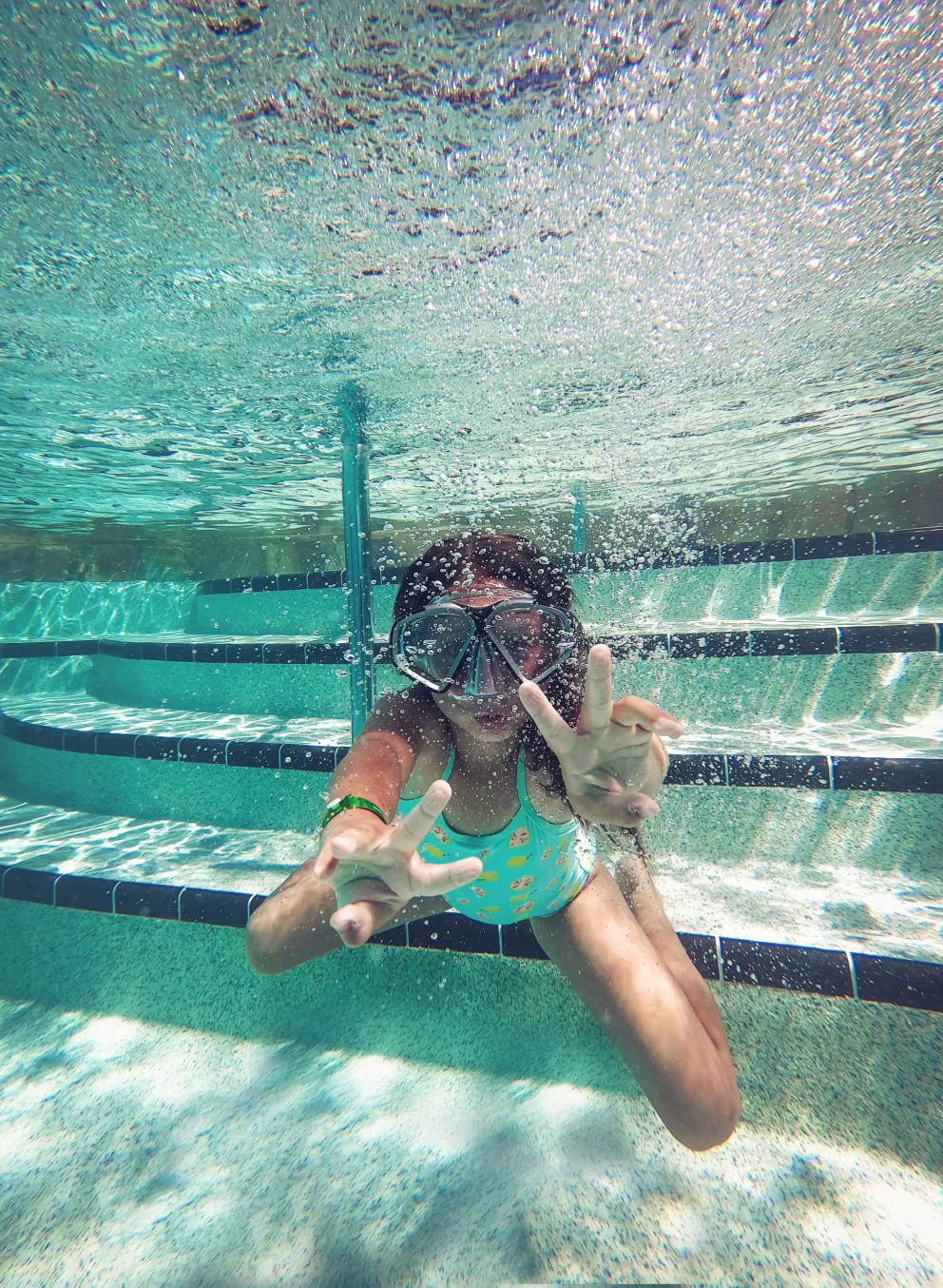 Child underwater in pool wearing snorkel mask, playfully forming peace signs with both hands.