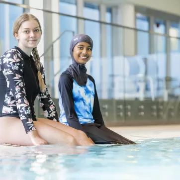 Two individuals in swimming gear sit on the ledge of an indoor pool with smiles on their faces.