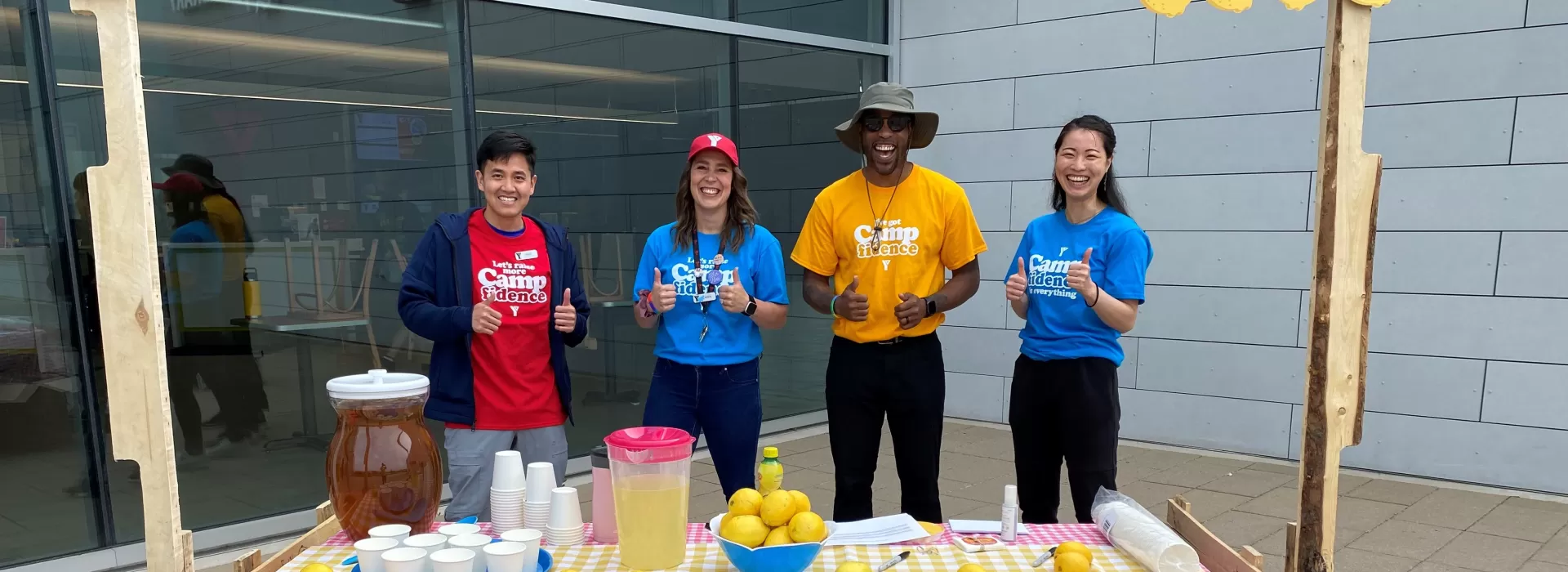 A group of 4 people stand in front of a wooden lemonade stand, smiling, in colourful t-shirts, to support the Camp It Forward campaign at YMCA Calgary.